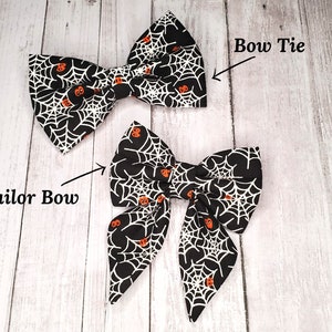 May include: Two black and white bow ties with a spiderweb and orange pumpkin pattern. The bow tie on the left is a sailor bow and the bow tie on the right is a bow tie.