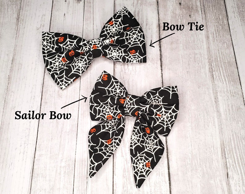 May include: Two black and white bow ties with a spiderweb and orange pumpkin pattern. The bow tie on the left is a sailor bow and the bow tie on the right is a bow tie.