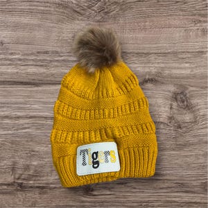 May include: Mustard yellow knit beanie with a faux fur pom-pom. The beanie features a white patch with the word "Tigers" in black and yellow lettering. The hat is made of a thick, textured knit.