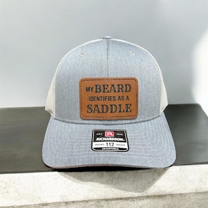 May include: A gray trucker hat with a white mesh back and a curved brim. The hat features a brown leather patch with the text "MY BEARD IDENTIFIES AS A SADDLE". The hat is adjustable and has the Richardson brand logo.