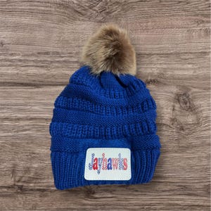 May include: A royal blue knit beanie with a faux fur pom-pom on top. The beanie features a white patch with the word "Jayhawks" in red, white, and blue lettering. The hat is made of a thick, textured material.