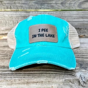 Puede incluir: Una gorra de camionero turquesa y beige con un parche marrón que dice "Meo en el lago".