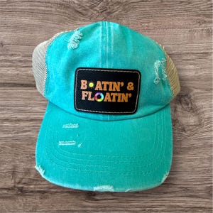 May include: A distressed turquoise trucker hat with a mesh back and a curved brim. A black patch on the front displays the text "BOATIN' & FLOATIN'" in orange and white. The hat has a worn, vintage look.