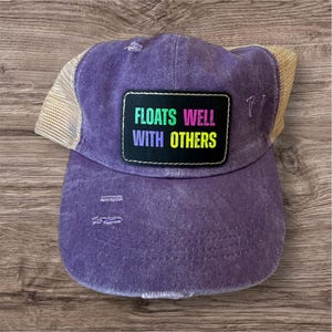 May include: A purple trucker hat with a distressed look. The front panel features a black rectangle with the text "FLOATS WELL WITH OTHERS" in colorful letters. The hat has a beige mesh back and a curved brim.