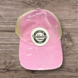 May include: Pink distressed baseball cap with a tan mesh back. The cap features a circular patch with the text "LET'S KEEP THE Dumbfuckery TO A MINIMUM TODAY". The hat has a curved brim and adjustable strap.