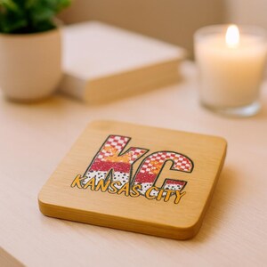 May include: A square wooden coaster with the letters "KC" in a colorful design, with the words "Kansas City" below. The coaster is on a light-colored surface, with a lit candle and a small plant in the background.