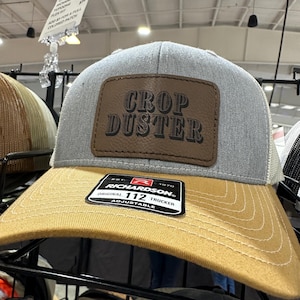May include: A gray and tan trucker hat with a brown leather patch that says "Crop Duster". The hat is adjustable and has the Richardson 112 logo on the patch.
