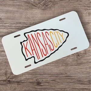 May include: White license plate with the words "KANSAS CITY" in red and yellow, outlined in black, inside a stylized arrowhead shape. The plate has four slots for mounting and is set against a wood grain background.