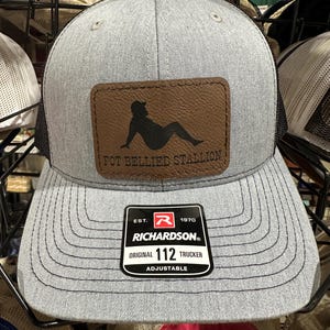 May include: A gray and black trucker hat with a brown leather patch that says "Pot Bellied Stallion". The patch features a silhouette of a person sitting down. The hat also has a black and red label that says "Richardson" and "Original 112 Trucker Adjustable".