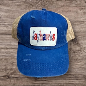 May include: A blue distressed baseball cap with a tan mesh back. The front features a white patch with the word "Jayhawks" in red, white, and blue checkered lettering. The cap has a curved brim and a distressed look.