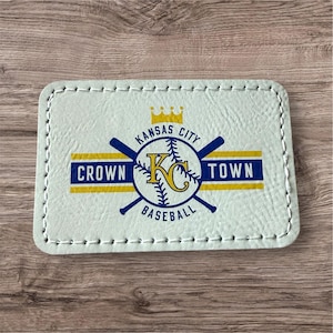 May include: A light green rectangular patch with white stitching around the edges. The patch features a baseball-themed design with the text "Kansas City Crown Town Baseball" in blue and yellow.