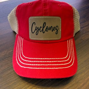 May include: Red and tan trucker hat with the word "Cyclones" on a brown patch. The hat has a curved brim with white stitching and a mesh back.