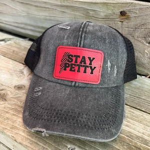 May include: Black distressed trucker hat with a red leather patch that says "STAY PETTY" in black letters with a checkered lightning bolt graphic.