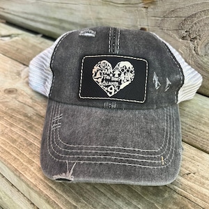 May include: A black and white distressed trucker hat with a white mesh back. The hat features a black leather patch with a heart design. The heart is filled with various Harry Potter themed icons and the text "Always" and "This Time It's Always".