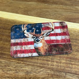 May include: Rectangular decorative item featuring a deer with large antlers set against a distressed American flag design. The flag has red and white stripes and a blue field with white stars. The deer is brown and white.