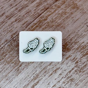 May include: A pair of stud earrings featuring a white wing design with green glitter accents. The earrings are displayed on a white card against a wooden background.