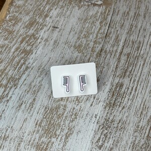 May include: A pair of comb-shaped earrings on a white card. The earrings feature a black comb design with a pink outline. The background is a light-colored, textured wooden surface.