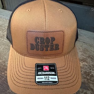 May include: A brown and black trucker hat with a brown leather patch that says "Crop Duster". The hat has a black mesh back and a red and black label that says "Richardson Original 112 Trucker Adjustable".