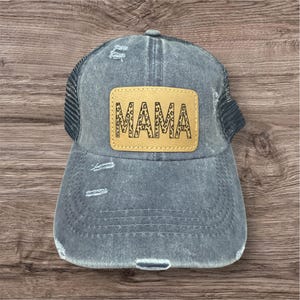 May include: A distressed denim baseball cap with a mesh back. The cap features a tan leather patch with the word "MAMA" in a leopard print design. The hat has a curved brim and is a faded blue color.