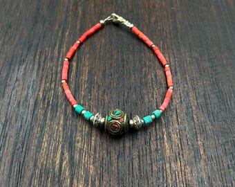 May include: A beaded bracelet featuring red and turquoise beads. The bracelet has a central decorative element with a gold and red design. The clasp is silver. The bracelet is displayed on a dark wood surface.