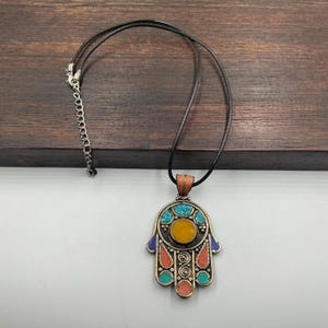 May include: A silver-toned Hamsa hand pendant necklace with a black cord. The pendant features a central yellow stone, surrounded by turquoise and coral-colored accents. The necklace has a silver chain clasp.
