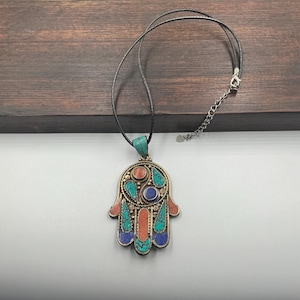 May include: A handcrafted pendant necklace featuring a Hamsa hand design. The pendant is adorned with turquoise, coral, and blue accents, set in a gold-toned metal. The necklace has a black cord and a silver-toned chain.