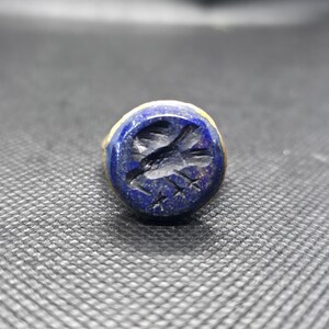 Könnte beinhalten: Ein runder, blauer Ring mit einem goldenen Band. Die Oberfläche des Rings zeigt ein geschnitztes Vogeldesign. Der Vogel ist dunkel vor dem blauen Hintergrund. Der Ring ist auf einer dunklen, strukturierten Oberfläche platziert.