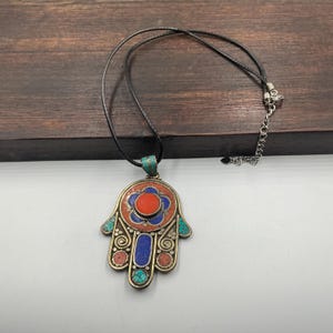 May include: A Hamsa hand pendant necklace with a black cord. The pendant features a detailed design with red, blue, and turquoise accents. The pendant is made of metal and has an intricate pattern.