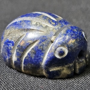 Op de afbeelding: Een gebeeldhouwde keverfiguur van lapis lazuli. De kever heeft een diepblauwe kleur met witte gebeeldhouwde details en een verweerde uitstraling. Het beeldje heeft een afgerond lichaam en een gedetailleerde kop met ogen en mond.
