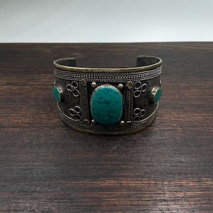 Può includere: Bracciale a polsino argentato con tre pietre color turchese. Il bracciale presenta un design intricato con elementi decorativi e una finitura strutturata. La pietra centrale è di forma ovale, affiancata da due pietre rotonde più piccole.