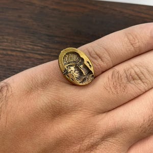 May include: Oval-shaped brass ring with a detailed relief of a figure wearing a helmet. The ring has a vintage, antique appearance, with a tarnished gold color. The design is centered on the ring's face, showcasing intricate details.