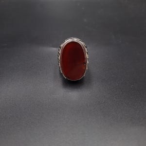 May include: A silver ring with an oval, reddish-brown stone. The ring's band has a decorative pattern around the stone. The ring is set against a dark gray background.