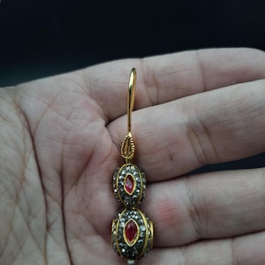 May include: Gold-toned earring with two stacked, beaded orbs. Each orb features a central red gemstone surrounded by small, sparkling accents. The earring's hook is also gold-toned, with a decorative loop at the top. The background is dark.