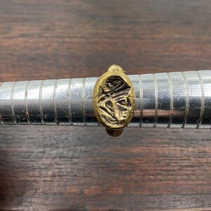 May include: A gold-colored ring with an oval face featuring a carved design of a face. The ring is displayed on a silver ring sizer. The ring has a vintage or antique appearance.