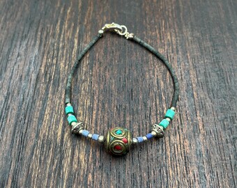 May include: A handmade bracelet featuring a central, ornate bead with red and green accents. The bracelet has turquoise and blue beads, and a black cord. The clasp is silver-toned. This piece is a unique accessory.
