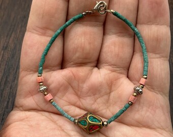 May include: A turquoise and coral beaded bracelet with a central geometric charm. The charm features red and turquoise accents. The bracelet has a silver clasp and small gold-colored beads. The bracelet is approximately 7 inches in length.