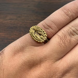 May include: A gold-colored ring with an oval-shaped face featuring a detailed religious figure. The ring has a textured, ornate border and is worn on a finger. The ring is made of metal.