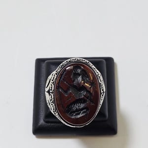 Könnte beinhalten: Ein Silberring mit einem ovalen, tief rotbraunen Stein, der ein dunkles, eingraviertes Intaglio-Motiv einer Figur mit Stab zeigt. Die Silberschiene weist auf den Seiten der Ringfassung komplizierte, dekorative Metallmuster auf.