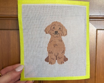 May include: Needlepoint canvas featuring a brown dog with a black nose and eyes. The dog is centered on a white mesh background, framed by a bright yellow border. The artwork is held by a hand with a gold ring.