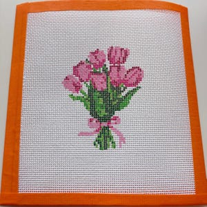 May include: Needlepoint canvas featuring a bouquet of pink tulips with green leaves and a pink ribbon. The floral design is set against a white background and framed by an orange border. A handmade floral design.