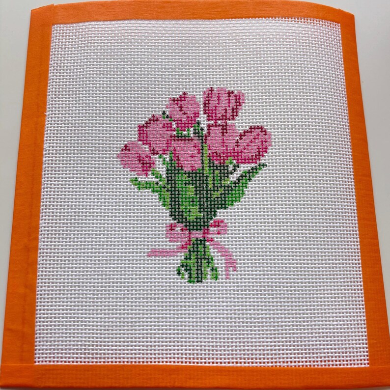 May include: Needlepoint canvas featuring a bouquet of pink tulips with green leaves and a pink ribbon. The floral design is set against a white background and framed by an orange border. A handmade floral design.
