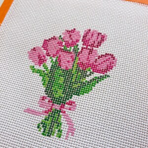May include: A cross-stitch design of a bouquet of pink tulips with green stems and leaves, tied with a pink ribbon. The needlepoint is on a white canvas.