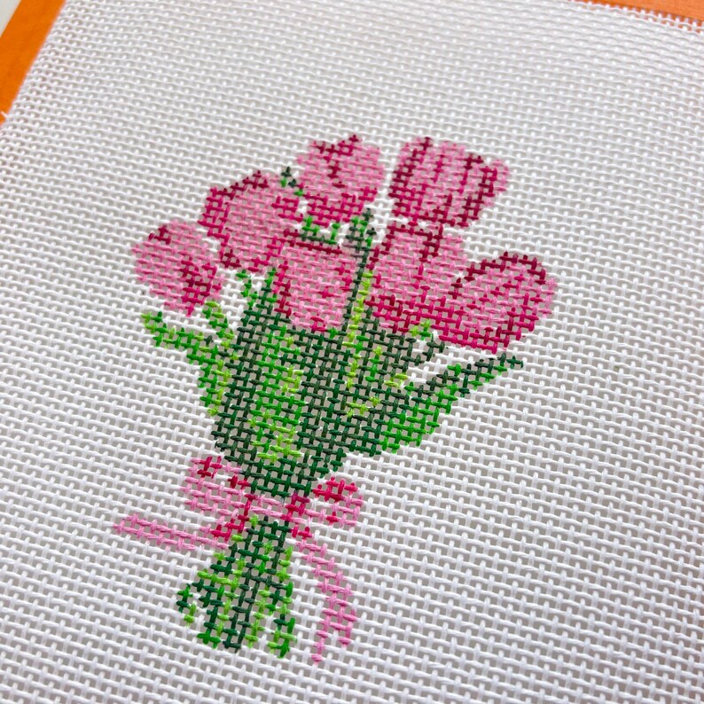 May include: A cross-stitch design of a bouquet of pink tulips with green stems and leaves, tied with a pink ribbon. The needlepoint is on a white canvas.