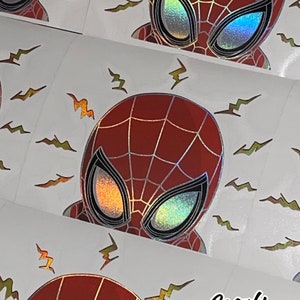 May include: A red and black holographic sticker featuring a cartoon image of Spider-Man with a red and black suit and a web design. The sticker has a white background with yellow lightning bolts.