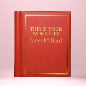May include: A red book with gold lettering and trim. The cover displays the text "THIS IS YOUR WORK LIFE" and "Janet Millard". The book is upright against a pink backdrop.