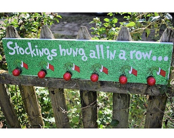 Puede incluir: Un letrero de madera verde brillante con el texto "Stockings hung all in a row..." presenta perillas rojas y obras de arte con temática de Grinch. El letrero está montado en una valla de madera rústica, con follaje verde en el fondo.