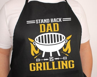 May include: Black apron with yellow text that reads "Stand Back Dad Is Grilling" with a white grill graphic and yellow flames.