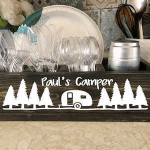 May include: A dark brown wooden box with a white camper and pine tree design. The box has a label that says "Paul's Camper". The box is filled with plastic utensils.