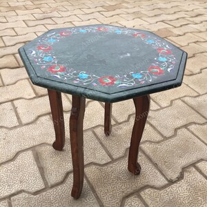 Puede incluir: Mesa octogonal de mármol verde con intrincados detalles florales y patas de madera. La mesa está sobre una superficie de ladrillo gris.
