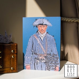 May include: An illustrated card featuring a man in a blue jacket and a white hat. The text on the card reads "Your face is my heart Sassenach and the love of you is my soul."